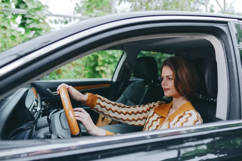 A woman driver sits in a car interior, hands on the steering wheel, calm expression, stylish sweater, natural daylight reflects on the scene.
