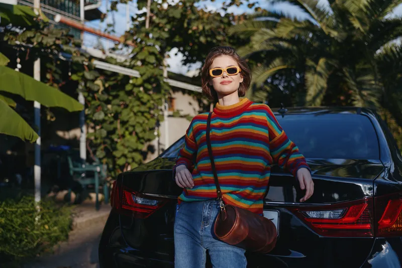 Woman in a colorful striped sweater and sunglasses leaning against a black car in a tropical outdoor street, casual outfit with jeans and leather crossbody bag, travel lifestyle and confident smile.