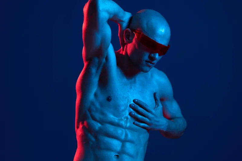 A strong man poses dramatically in a vibrant blue and red lighting environment, showcasing his toned physique. The image has a striking, modern aesthetic.