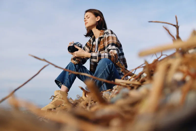 Photographer, camera, outdoor, beach, nature, woman, scenery, coastal, driftwood, casual woman crouches among weathered driftwood to shoot rugged shoreline with focused expression