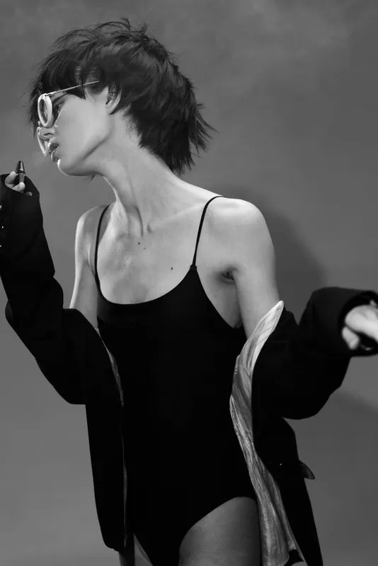 Black and white portrait of a woman wearing sunglasses and a black bodysuit with a loose jacket, exuding confidence and style.