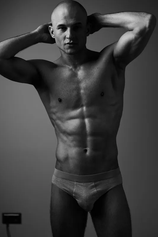 Black and white portrait of a muscular man posing confidently, showcasing his toned physique and body language in a minimalist setting.