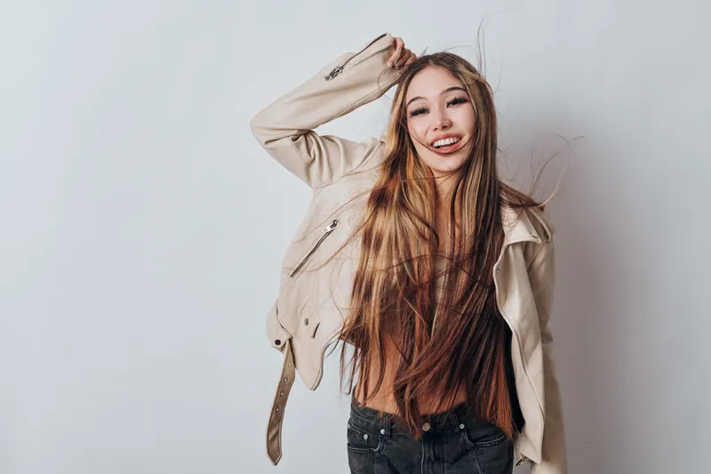 Young woman with long hair wearing beige jacket smiling and posing against light background in studio. Casual style, happy female model with natural look and loose hair movement.