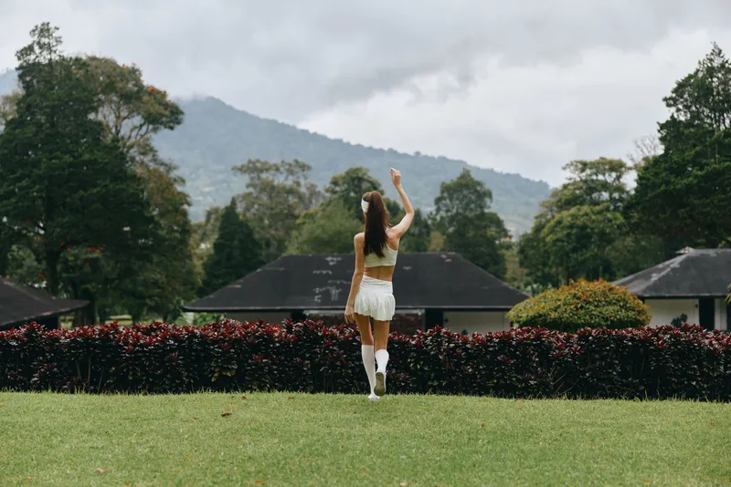 Young woman in white sportswear waving, standing in a lush green landscape with mountains in the background, conveying joy and fitness