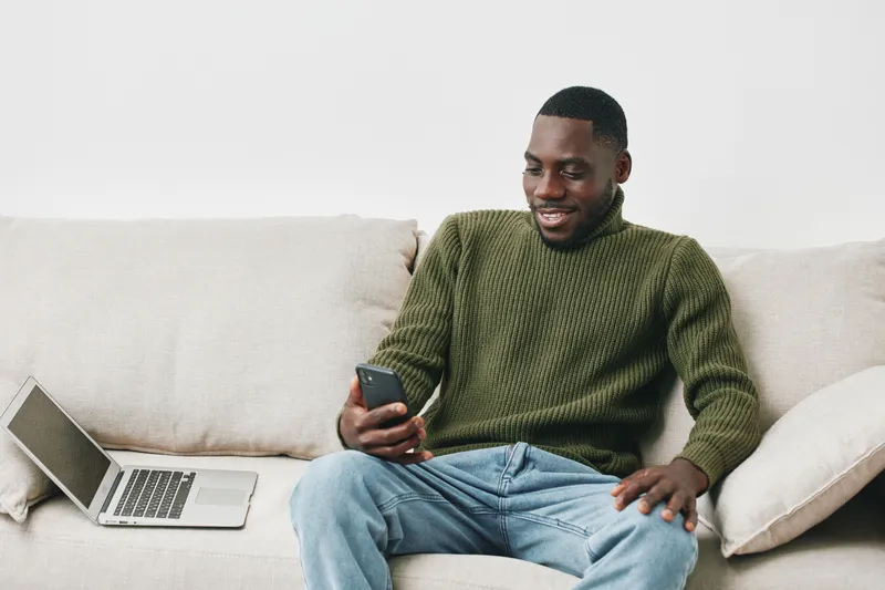 Young Black man enjoying time on smartphone while sitting on couch with laptop, showcasing relaxed lifestyle and modern technology