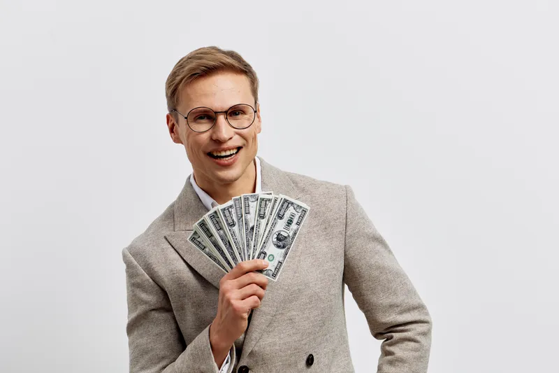 Happy young man wearing glasses holds a fan of hundred-dollar bills and smiles confidently, dressed in a beige blazer. Studio shot on plain white background, finance success concept, business lifestyl