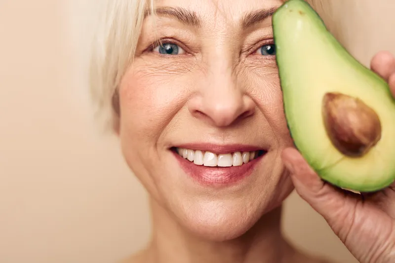 Smiling mature woman with clear skin holds half an avocado near her face on beige background. Concept of healthy lifestyle, natural beauty, skincare, nutrition, and wellness.