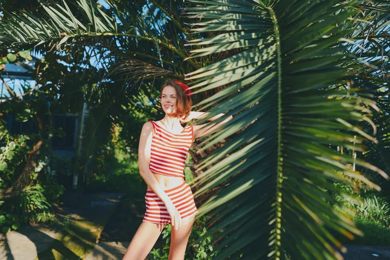 A cheerful woman in a striped red and white swimsuit stands among tropical foliage, basking in sunlight. She smiles brightly, exuding confidence and carefree energy in a vibrant outdoor setting.