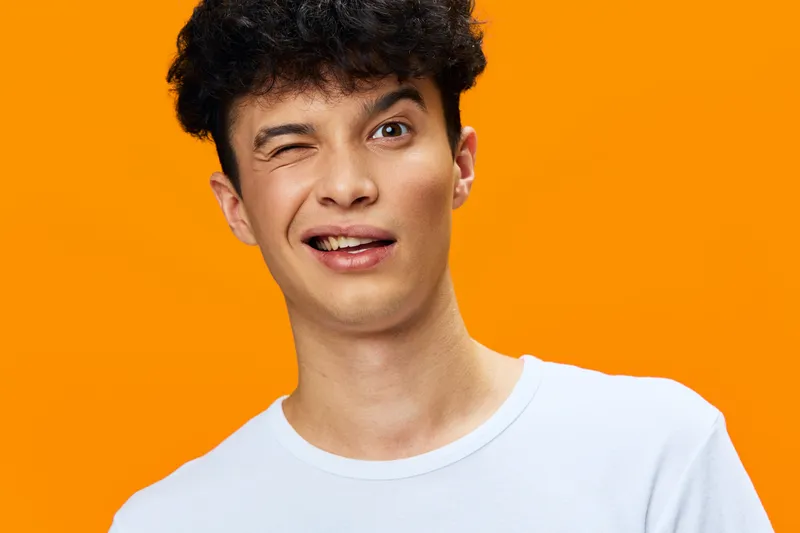 Young man winking playfully against orange background wearing casual white t-shirt, expressing fun and lighthearted mood with curly hair and bright studio lighting.