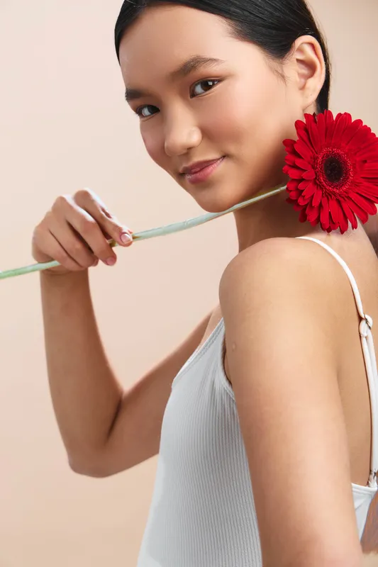 asian woman holds a bright red flower near her shoulder in a soft studio setting. She wears a light knit top and smiles gently, exuding elegance and calm.
