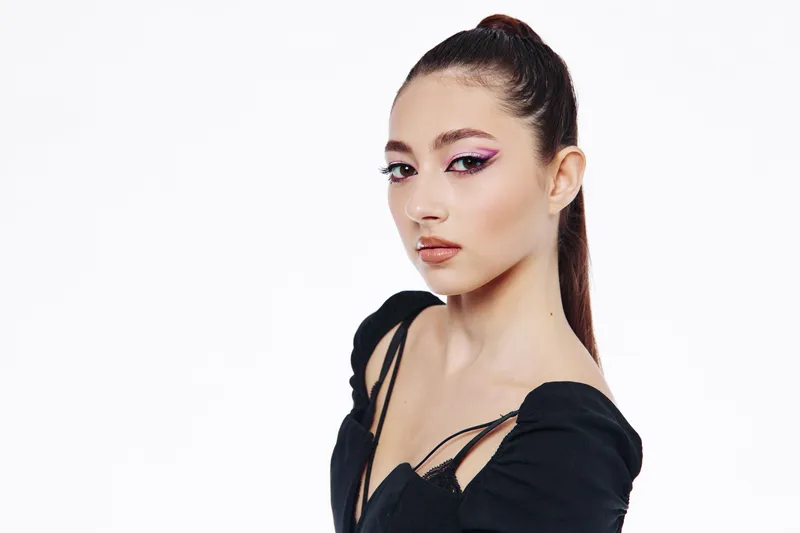 Fashion model with sleek ponytail and bold purple eyeliner poses confidently wearing black high fashion garment on clear white background. Beauty trend for editorial style.