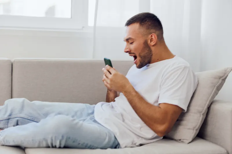 A surprised man engaging with his smartphone, expressing emotions in a cozy living room, reflecting on modern tech connection and personal space.