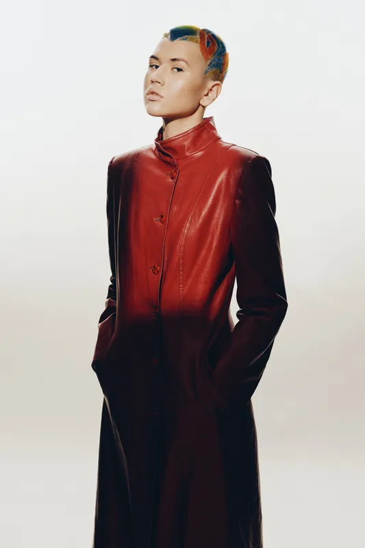 Woman with colorful short hair wearing a red leather coat, standing confidently with hands in pockets against a plain light background, modern fashion style.