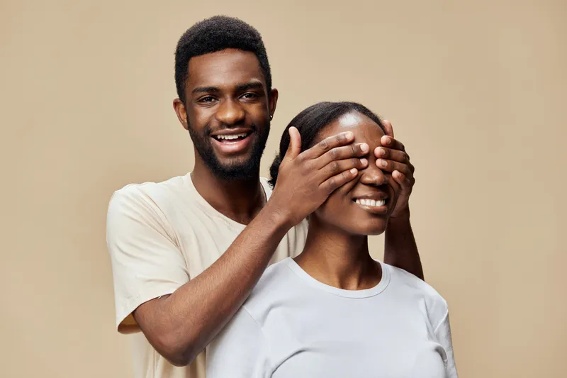 Happy couple playfully interacting with one partner covering the other s eyes, expressing joy, featuring a neutral beige background