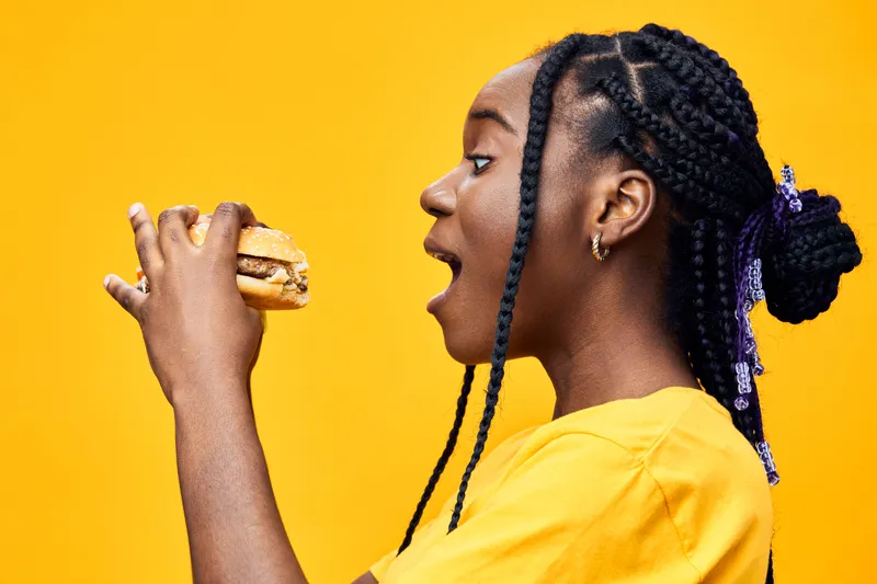 Young woman with braids excitedly holding a burger against a vibrant yellow background, showcasing a joyful moment with delicious food