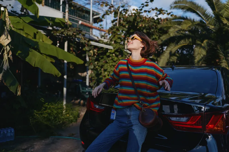 Woman in her 20s wearing a colorful striped sweater and sunglasses, leaning on a black car trunk by palm trees, smiling and looking up in a casual travel lifestyle scene.