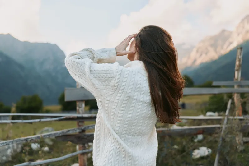 Smiling woman wearing a white knit sweater standing in a mountain meadow back turned flowing hair soft natural light relaxed outdoor lifestyle mood