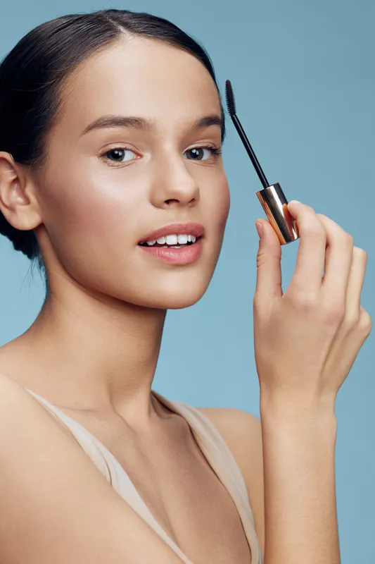 Young woman applying mascara, showcasing beauty and confidence against a soft blue background, highlighting makeup and skincare concepts