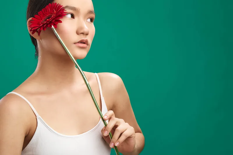 asian woman is photographed in a studio against a teal backdrop, holding a bright red flower near her face. a calm, elegant portrait with soft lighting conveys grace and a thoughtful, serene mood.