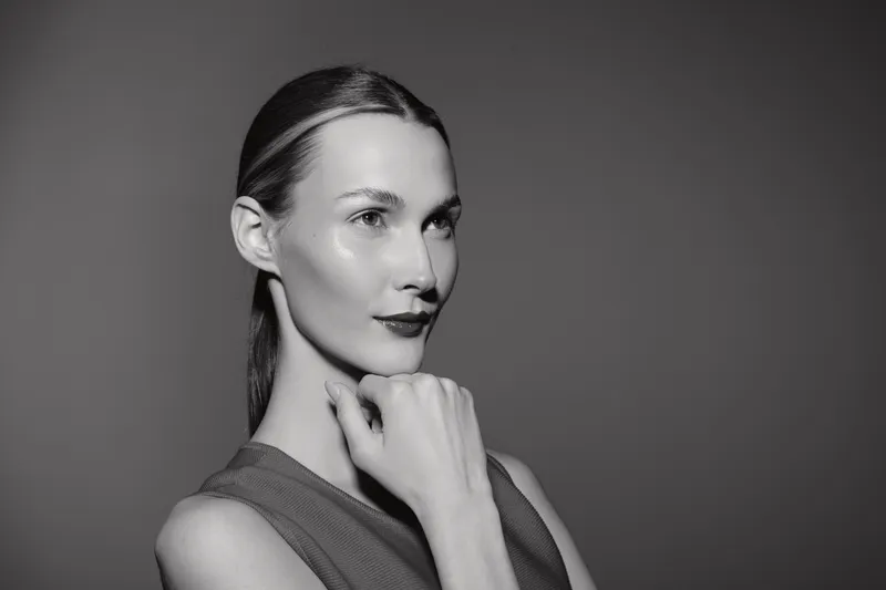 Portrait of a slender white woman with fair skin and slicked back hair, posing thoughtfully against a grey background.