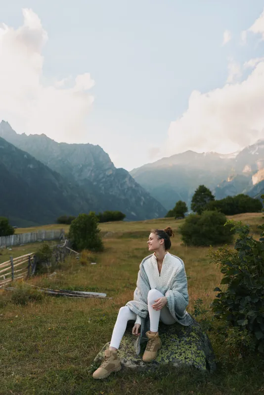 Young woman sits on a rock in a sunlit meadow with mountain backdrop, enjoying a peaceful moment outdoors.