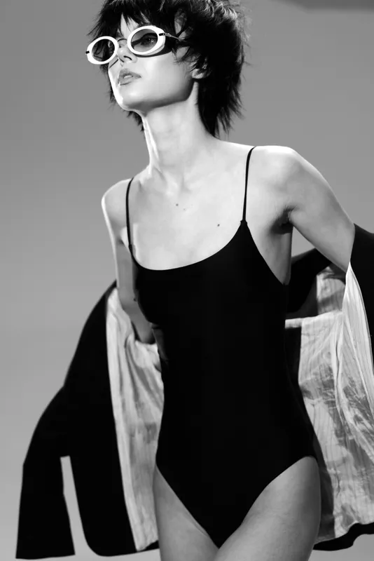 Black and white portrait of a stylish woman in sunglasses wearing a black swimsuit with a loose jacket on a gray background.