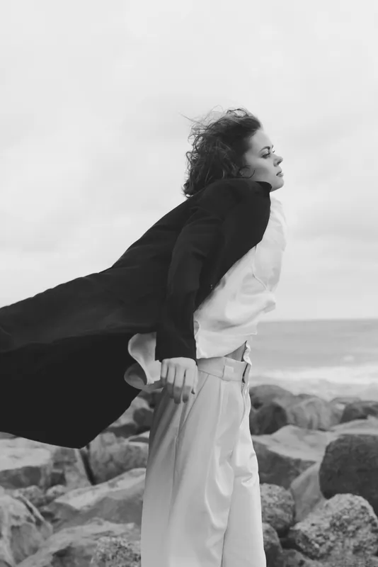 Wind swept cape and flowing garments by the ocean, a dramatic fashion moment in a coastal monochrome scene, capturing movement, contrast and moody editorial styling against a rocky shoreline