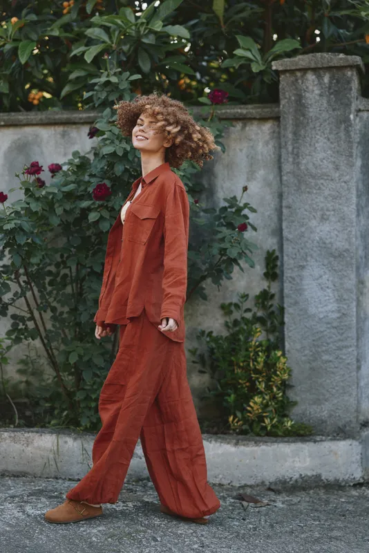 A stylish woman walks along a city street in a rust colored jumpsuit, exuding confidence and calm. Natural light highlights casual elegance and lifestyle chic in an urban moment