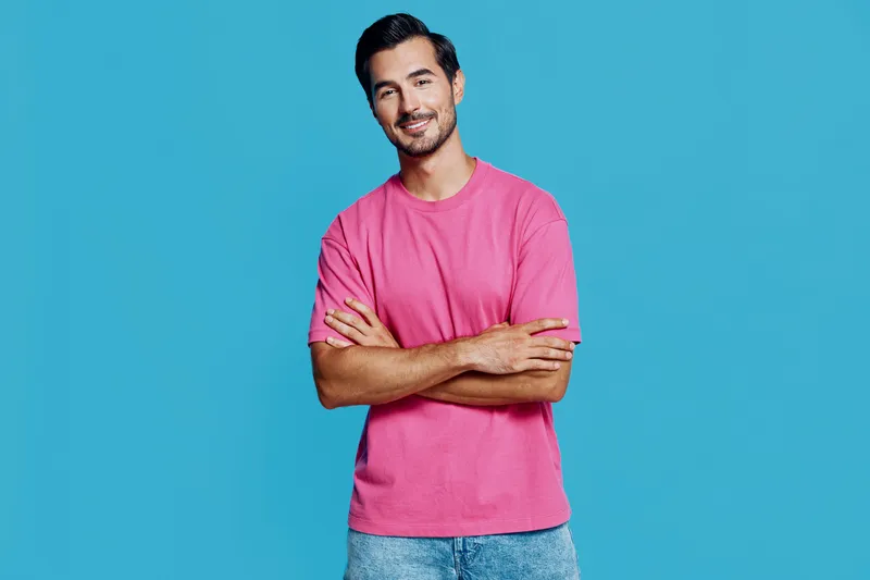 Smiling man wearing a pink t-shirt, standing against a vibrant blue background with arms crossed, exuding confidence and happiness in a cheerful portrait.