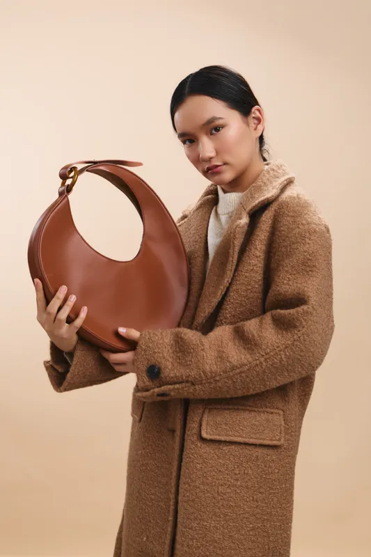 asian woman in a warm beige studio poses with a stylish brown bag, exuding calm confidence and fashion sense, capturing a modern urban lifestyle moment.