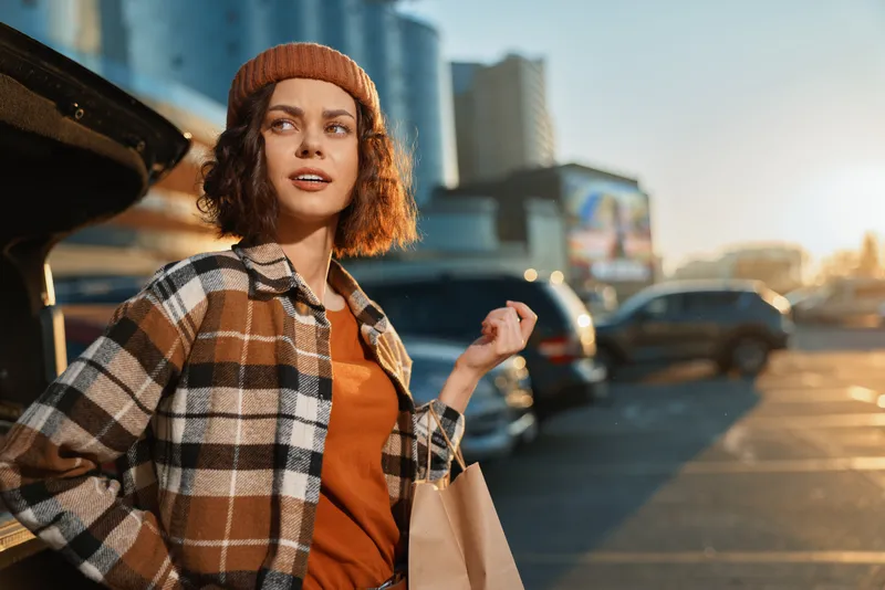 Woman shopping in urban parking lot at golden hour, candid portrait conveying authenticity and lifestyle, emotional smile in autumn plaid jacket and beanie with mindful living vibe and storytelling.
