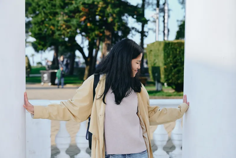 Woman in a beige coat leaning between white columns outdoors, showcasing casual fashion and relaxed pose in a garden setting with greenery and architectural backdrop