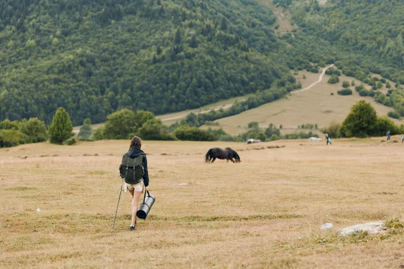 A lone hiker with a backpack walks across a vast grassy field toward distant hills, capturing a serene outdoor moment in a tranquil rural landscape.