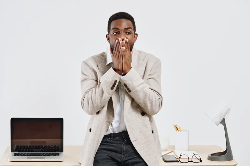 Close-up of surprised young African American man wearing beige blazer and white shirt, covering mouth with hands, looking at camera, indoor office background with laptop, lamp, and stationery, human e