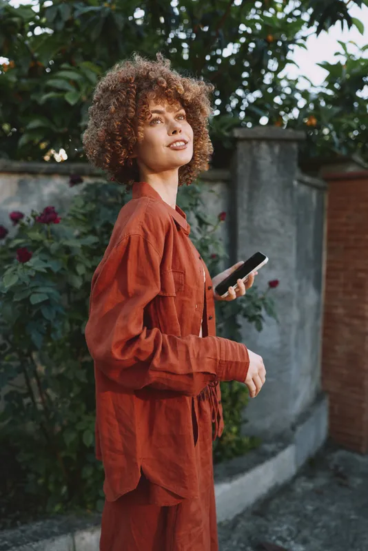 Woman lifestyle portrait outdoors in rust red outfit holding smartphone, natural light, confident pose, urban garden backdrop, candid moment