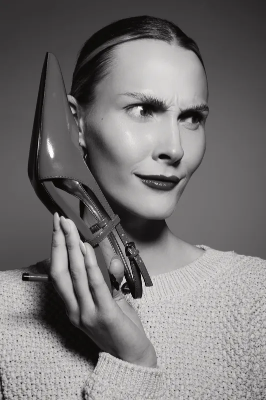 A white woman with fair skin holds a high heel shoe near her face with a puzzled expression. Black and white portrait with moody lighting, fashion and beauty concept.