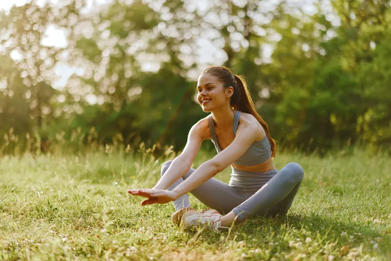 Young athletic woman in a light grey sports outfit stretching outdoors, expressing joy amidst greenery, perfect for fitness and healthy lifestyle themes