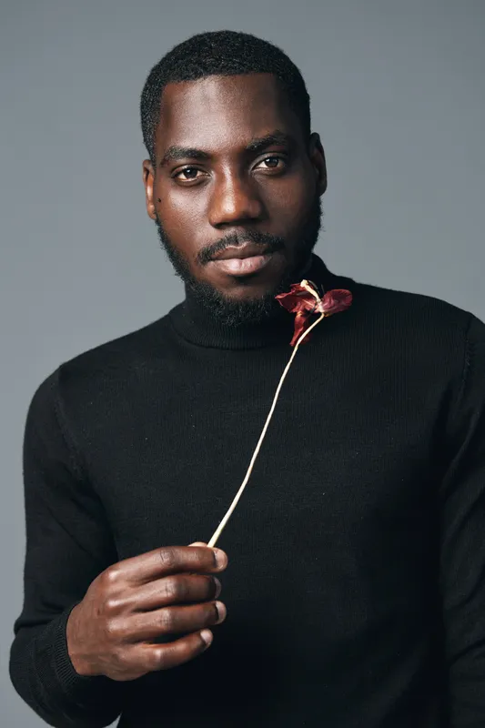 African man with dark skin holding dried flower in hand wearing black turtleneck sweater in creative conceptual visual style portrait with serious expression on gray background.