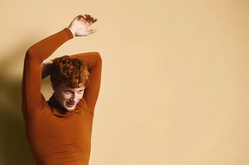 Woman with short red hair wearing orange sweater posing with hands above head standing in front of beige background. Expression shows calm and relaxed mood in studio.