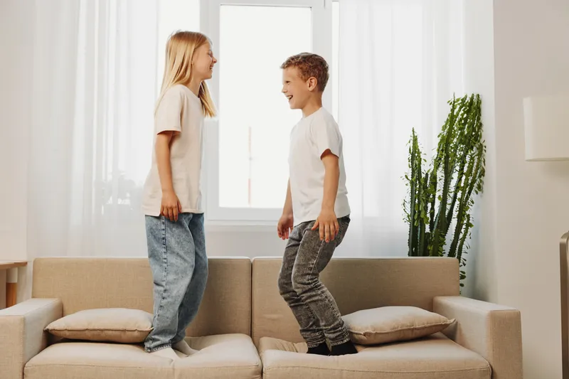 Two happy children jump on a beige sofa in a bright living room with natural light and green plants, enjoying playful moments and family fun indoors.