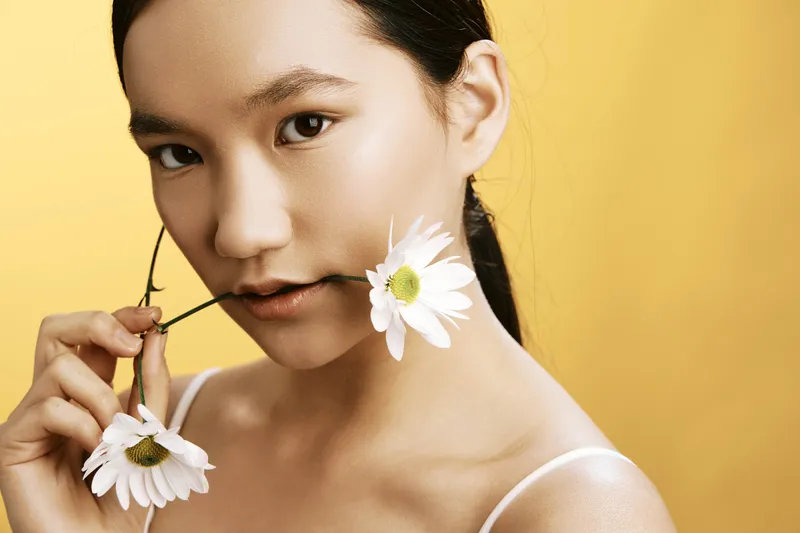 Asian woman with flowers near her lips stands in a vivid studio portrait, radiating grace and calm with a natural, elegant look and minimal, warm background.