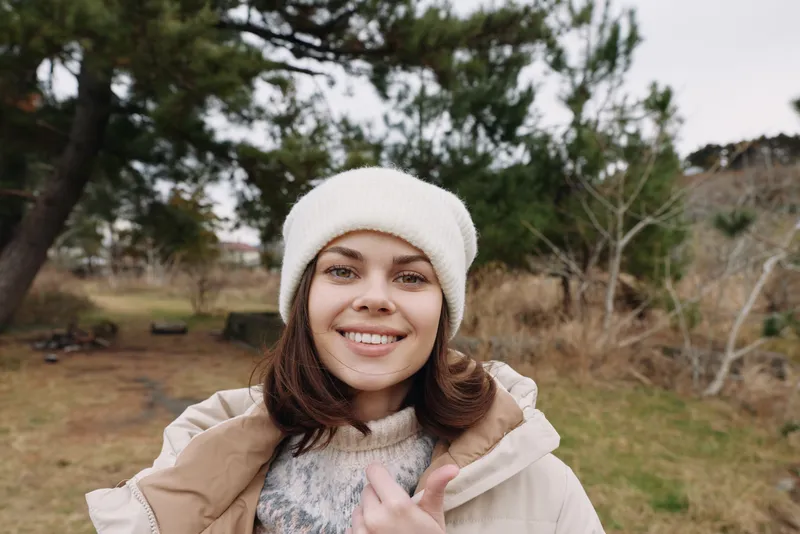 Woman outdoor portrait in winter attire, smiling in a park setting with a cozy coat and knit hat, capturing candid fashion and a relaxed mood under natural daylight.