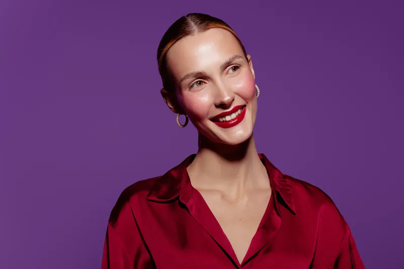 Smiling woman with red lipstick and hoop earrings wears red satin blouse on purple background. Portrait of confident female model with natural makeup and elegant style.