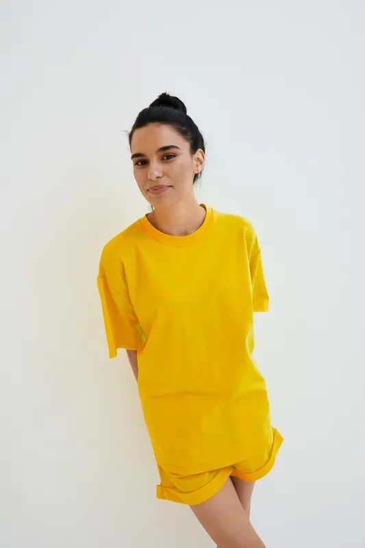 Young woman wearing bright yellow casual clothes stands against white background with relaxed expression, natural makeup, and tied-back hair in minimalistic studio setting.