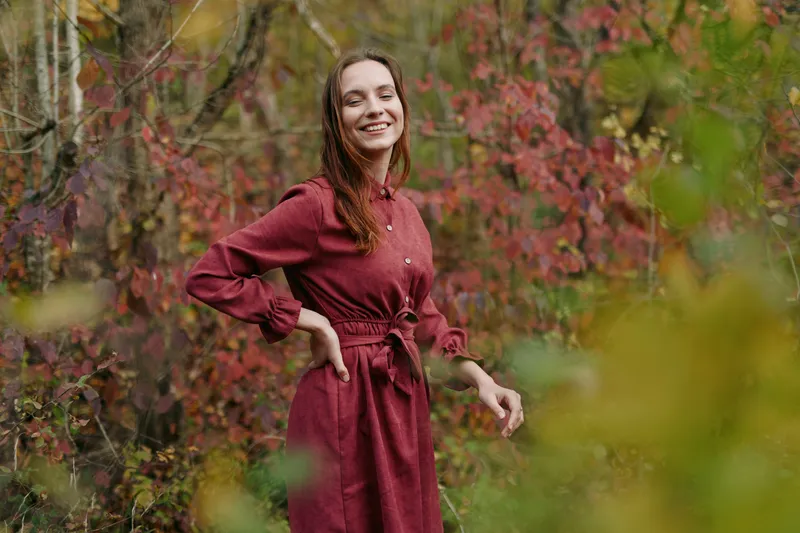 A candid portrait of a smiling young woman in a burgundy dress among autumn foliage, conveying authenticity and credibility through a natural pose, warm light and a genuine expression.