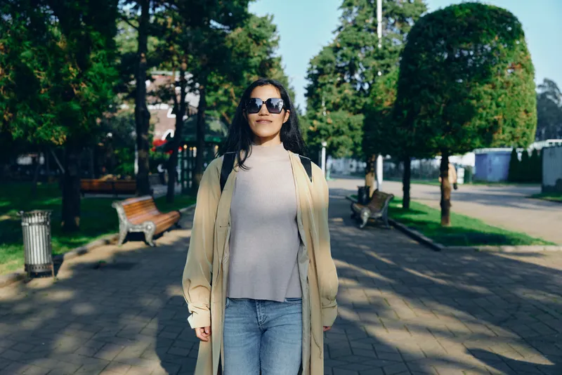 Woman in a long beige coat strolling through a sunny park, stylish sunglasses, casual chic mood, outdoor urban scene, relaxed daytime fashion