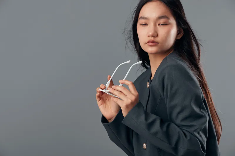 Young woman in dark blazer holding white eyeglasses with long straight hair looking down, studio portrait on gray background. Casual fashion, confident female model with minimal makeup and natural