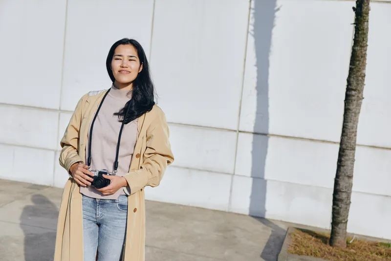 Woman holding camera outdoors in a light trench coat and denim, standing beside a white wall with a tree, capturing everyday moments in street style, casual lifestyle photography