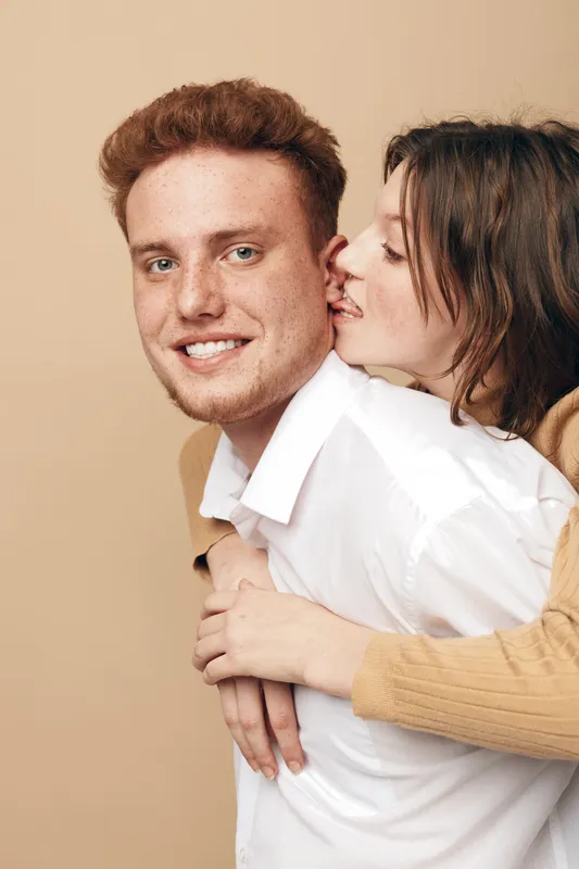 Young couple in casual clothes share intimate moment, woman hugging man from behind and whispering in his ear. Concept of love, affection, relationship, and happiness.