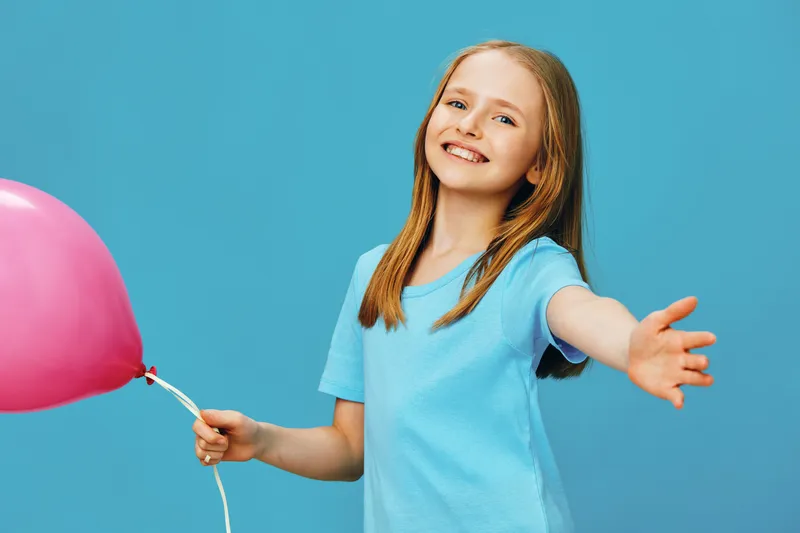 Happy young girl with a pink balloon, smiling brightly against a vibrant blue background, embodying joy and playfulness in a colorful, cheerful portrait